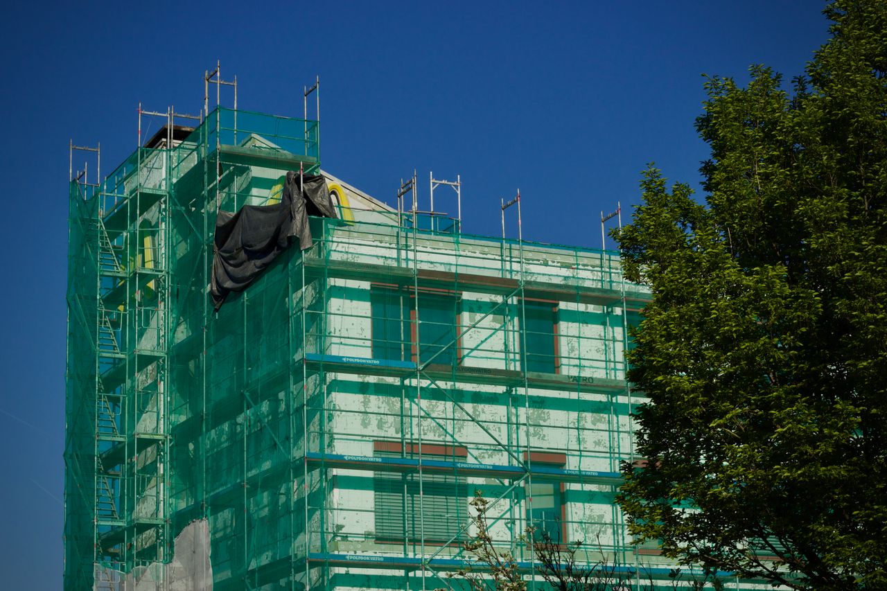 home-services-02 Building under renovation covered with green scaffolding against a clear blue sky.
